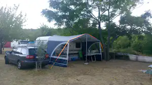 Großes Campingzelt mit Vorzelt, daneben ein Auto und ein Tisch auf dem Stellplatz, Bäume im Hintergrund.
