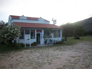 Campinghaus mit Terrasse, Eingangstür und Fenstern, davor Kiesweg und Rasenfläche, Bäume und Hecken am Rand.