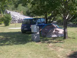 Parkende Autos auf einem Campingplatz im Grünen, neben einem Zelt und unter Bäumen, mit Steinschüttung im Hintergrund.