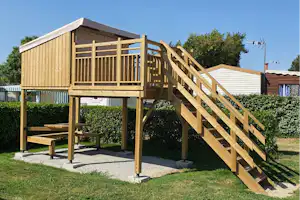 Holzspielhaus auf Stelzen mit Treppe, Sitzbank und Rasenfläche auf einem Campingplatz, im Hintergrund Hecken und Gebäude.