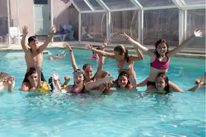 Familie im Schwimmbecken eines Campingplatzes, Kinder und Erwachsene im Wasser vor einer überdachten Poolanlage.