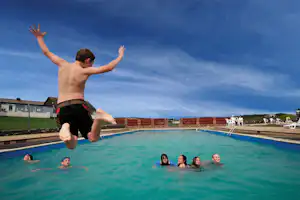 Familie springt ins Schwimmbecken eines Campingplatzes, Kinder im Wasser und Sprung eines Jungen vor blauem Himmel.