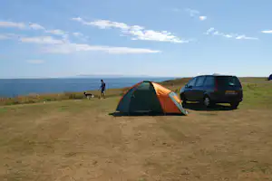Orangefarbenes Campingzelt auf einer Wiese am Meer, daneben ein Auto und ein Hund, im Hintergrund Küste und Hügel.