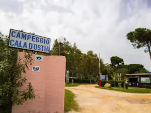 Camping-Eingang mit Schild „Campeggio Cala d’Ostia“ und „Bar“, Weg und Hecken im Außenbereich.
