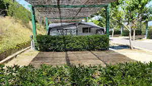 Wohnwagen auf einem Campingplatz mit Holzterrasse, Hecke und Bäumen im Vordergrund, überdachter Stellplatz im Hintergrund.