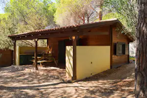 Holzbungalow mit Terrasse, Tisch und Grillbereich, umgeben von Bäumen und Kiesweg am Campingplatz.