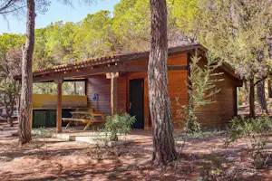 Holzhütte mit überdachter Veranda, Tisch und Grillbereich, umgeben von Kiefern und mediterraner Vegetation im Campingwald.