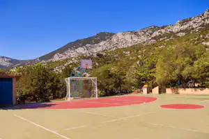 Sportplatz mit Basketballkorb auf rotem Feld, umgeben von Bäumen und Bergen im Hintergrund.