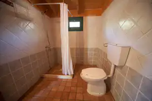 Toilette im Sanitärraum mit weißem WC, Duschvorhang, Fliesenboden und Wandfliesen, Holzdecke und Handbrause an der Wand.