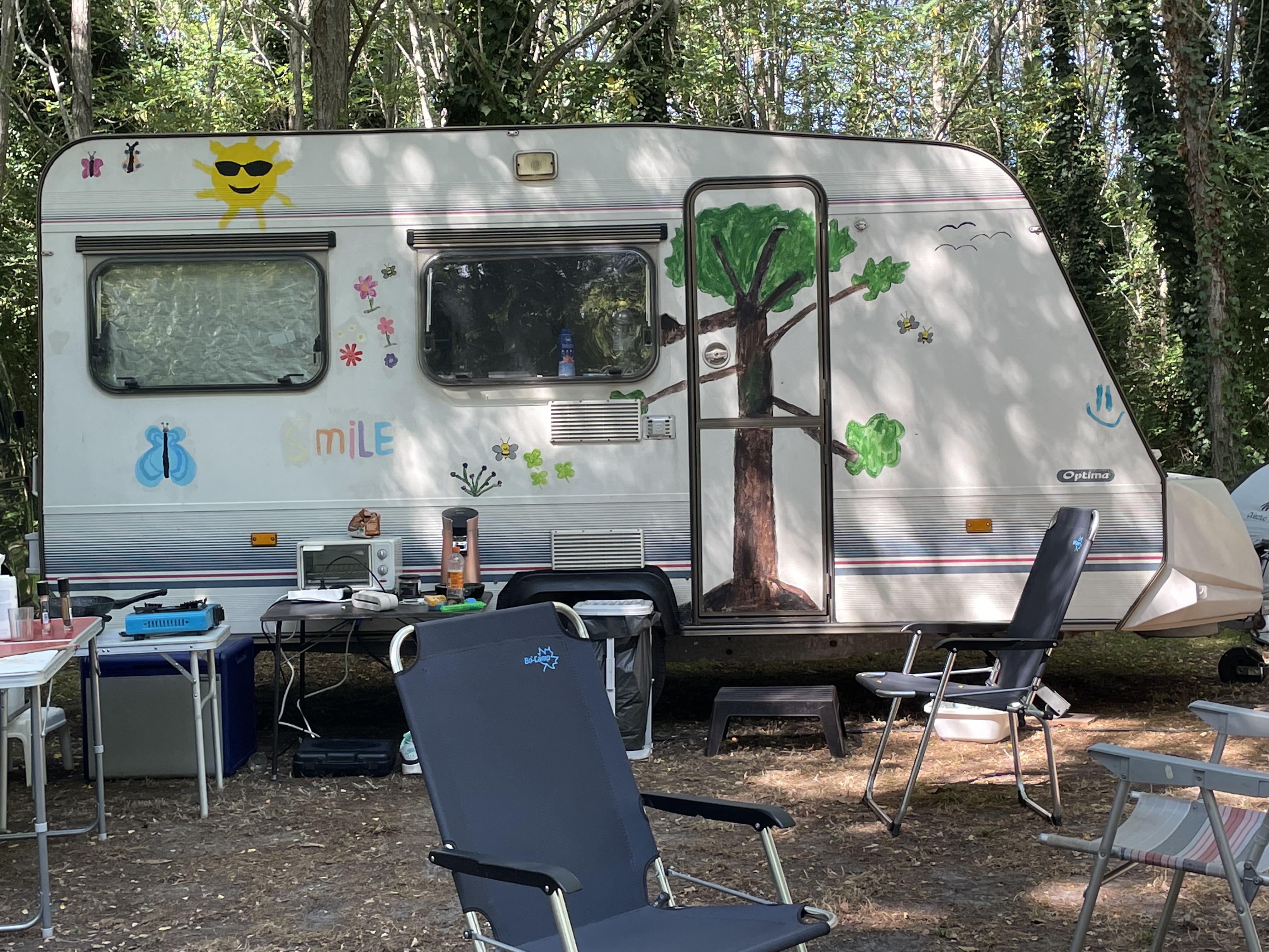 Wohnwagen mit Vorzelt und Terrasse, Campingstühle und Tisch davor, umgeben von Bäumen im Wald.