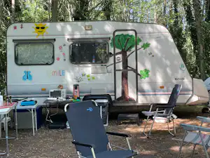 Wohnwagen mit Vorzelt und Terrasse, Campingstühle und Tisch davor, umgeben von Bäumen im Wald.