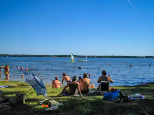 Camping am See: Badegäste sitzen am Ufer auf Decken, im Wasser schwimmen Menschen, Segelboote sind zu sehen.