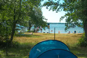 Zelt auf einem Campingplatz am See, im Hintergrund Uferpromenade und Bäume, Liegestühle am Strandbereich.