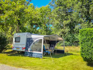 Wohnwagen mit Vorzelt auf einem Campingplatz, umgeben von Rasen und hohen Bäumen bei blauem Himmel.