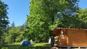 Blaues Zelt neben einer Holzunterkunft auf einem Campingplatz im Grünen, umgeben von Bäumen und Wiese.