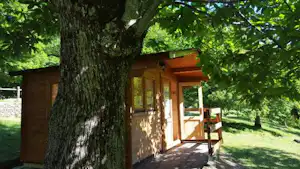 Holzhütte am Campingplatz mit Veranda und Sitzbank, umgeben von Bäumen und Rasen im Grünen.