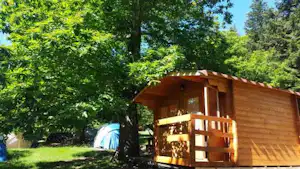 Holzsauna mit Terrasse am Campingplatz im Grünen, daneben ein Zelt zwischen Bäumen und Rasen.