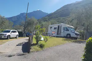 Wohnmobil auf einem Stellplatz mit Kiesfläche, daneben ein Auto, Hecken und Berge im Hintergrund.