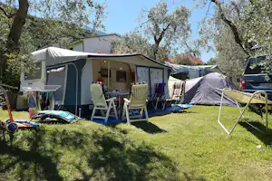 Campingplatz mit Wohnwagen und Zelt, davor Stühle und Tisch auf grünem Rasen, Bäume im Hintergrund.