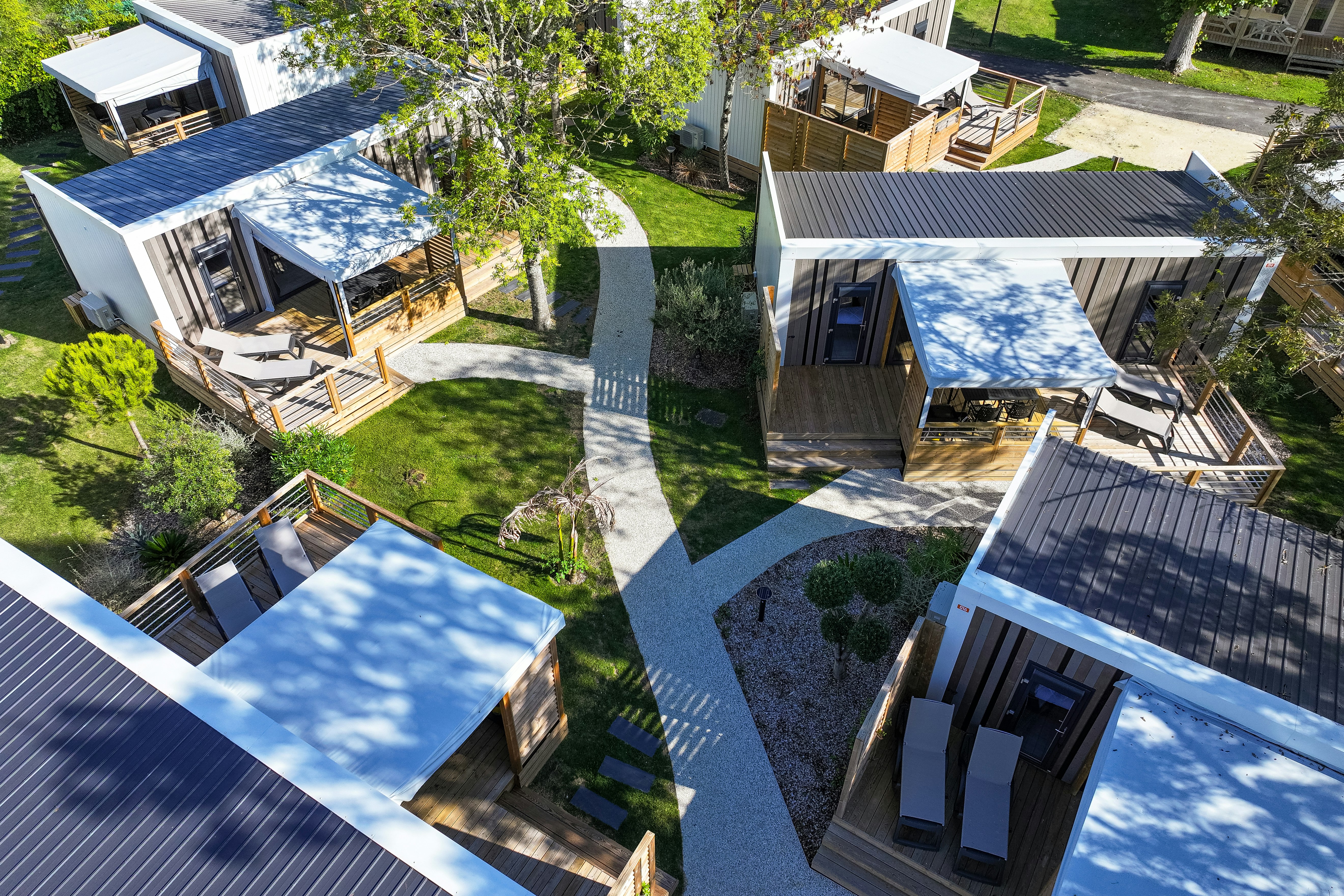 Luftaufnahme von mehreren Ferienhäusern mit Terrassen, Sonnenliegen und Gartenwegen, umgeben von Bäumen und Rasenflächen.