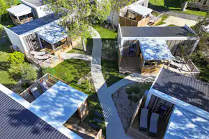 Luftaufnahme von mehreren Ferienhäusern mit Terrassen, Sonnenliegen und Gartenwegen, umgeben von Bäumen und Rasenflächen.