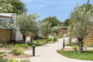 Campingweg zwischen Wohncontainern, mit Kiesbeeten, Rasen und Bäumen, im Hintergrund Gebäude und Holzterrassen.