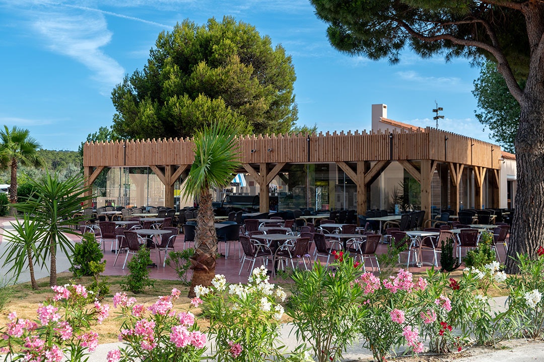 Terrasse mit vielen Stühlen und Tischen, Holzzaun und Bäumen, im Vordergrund blühende Blumen im Campingbereich.