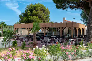 Terrasse mit vielen Stühlen und Tischen, Holzzaun und Bäumen, im Vordergrund blühende Blumen im Campingbereich.