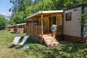 Mobilheim mit Holzterrasse und Stufen, daneben Liegestühle auf dem Rasen, umgeben von Bäumen und Bergen im Hintergrund.
