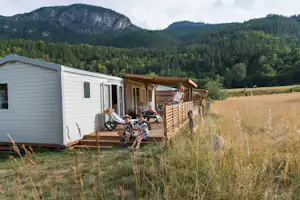 Wohnwagen mit Holzterrasse und Sitzgruppe, davor Gras und hohe Wiesen, im Hintergrund bewaldete Berge.