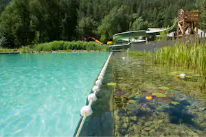 Türkisfarbenes Schwimmbecken am See mit Steg und Leiter, im Hintergrund Wald und ein Holzsteg am Ufer.