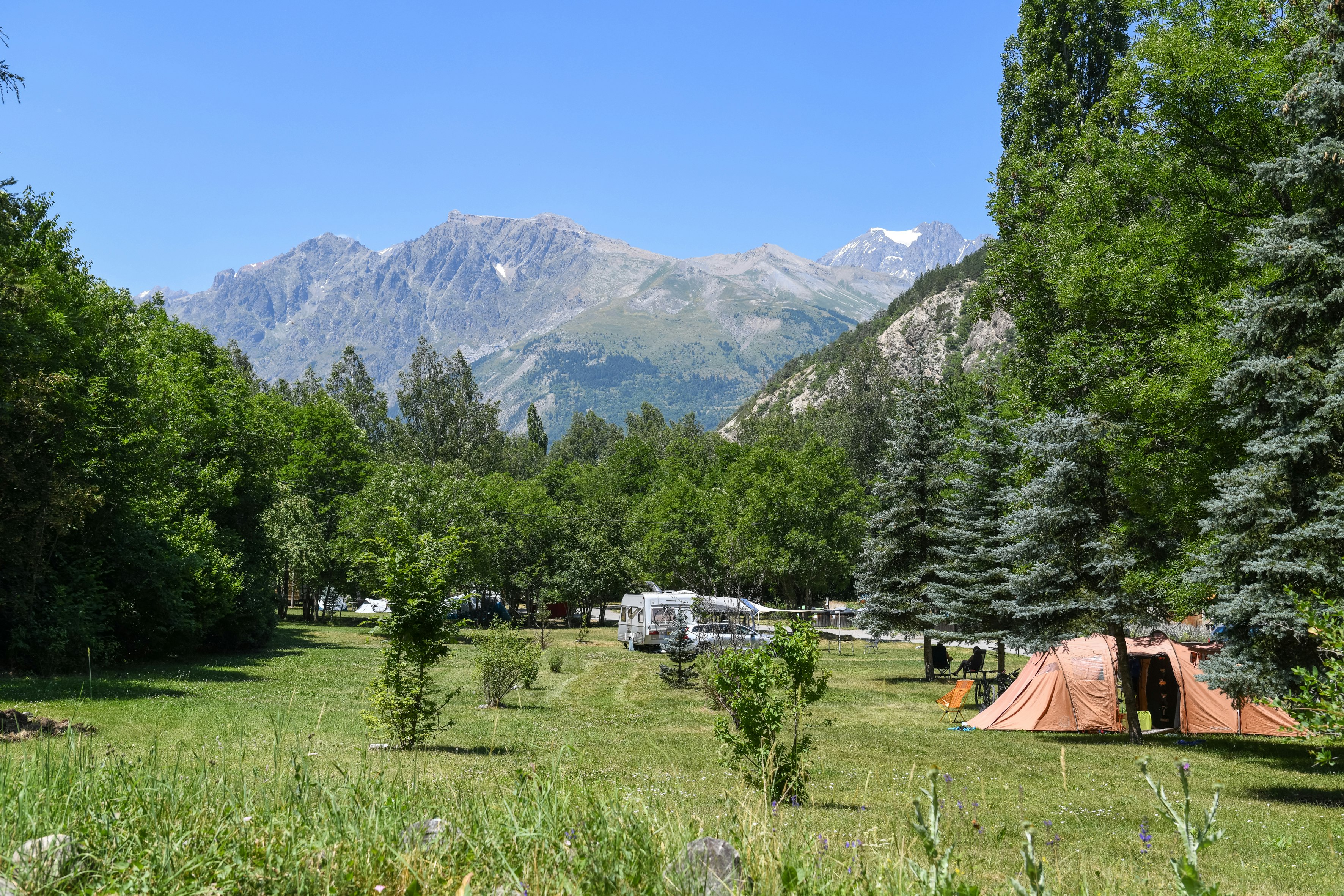 Campingwiese mit Zelt, Wohnwagen und Bäumen vor hohen Bergen im Hintergrund.