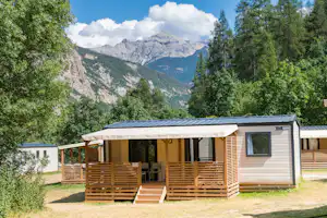 Modernes Camping-Mobilheim mit Holzveranda und Stufen, umgeben von Bäumen, im Hintergrund hohe Berge und Wolken.