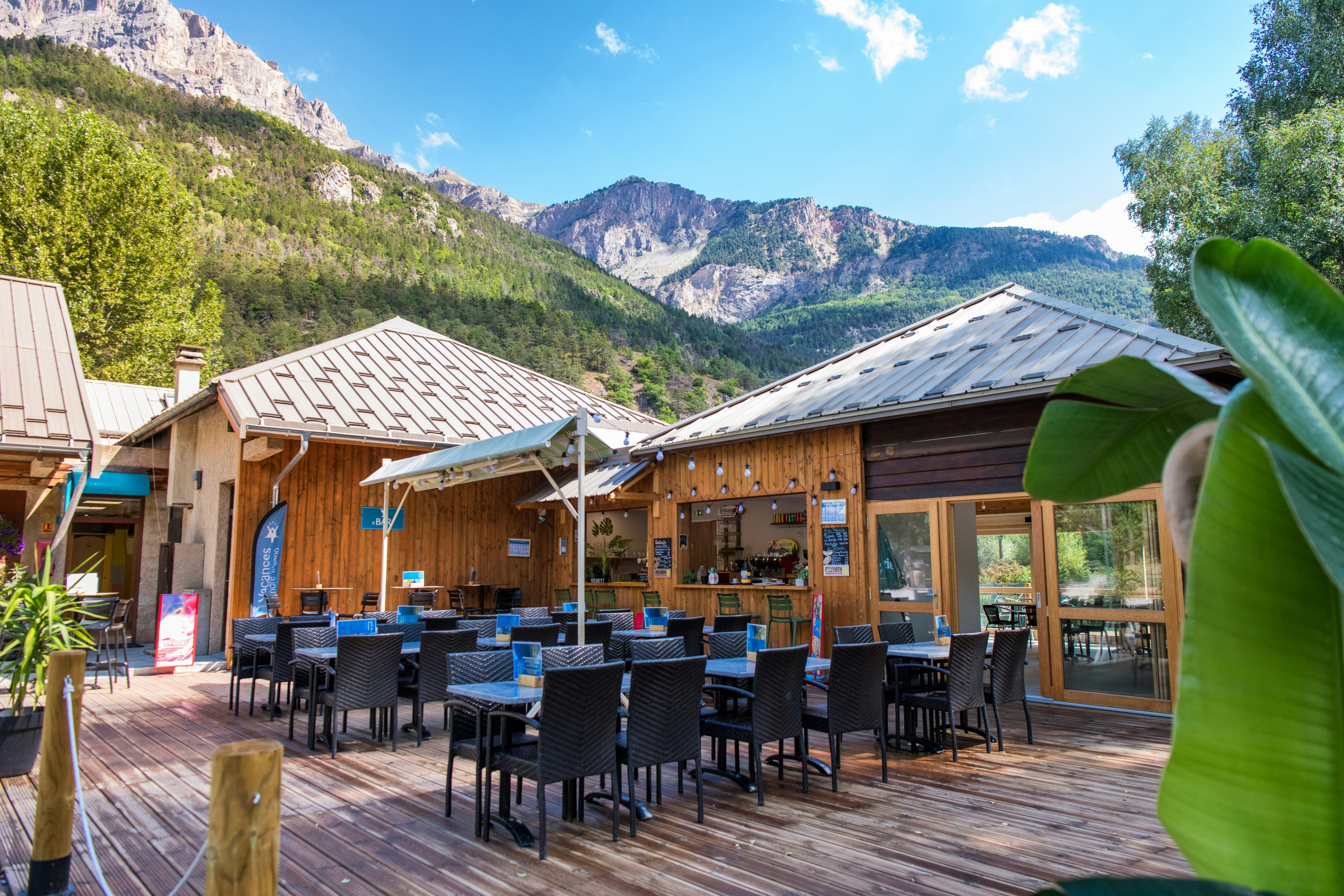 Holzterrasse mit vielen schwarzen Stühlen und Tischen, überdachte Bereiche und Zelt, umgeben von Bergen und grünen Bäumen.