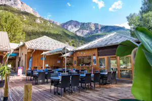 Holzterrasse mit vielen schwarzen Stühlen und Tischen, überdachte Bereiche und Zelt, umgeben von Bergen und grünen Bäumen.