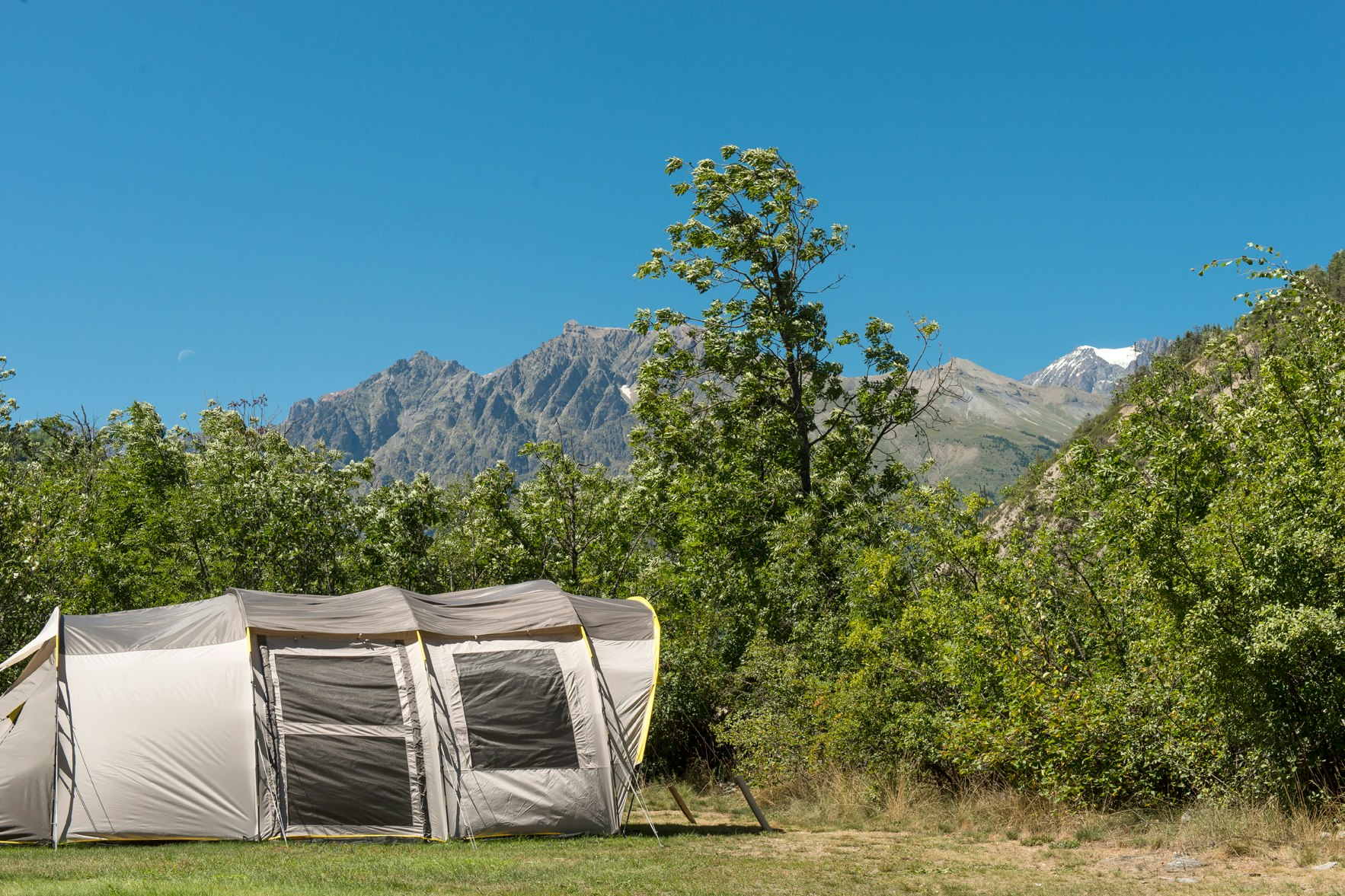 Zelt auf einem Campingplatz mit Bäumen und Bergen im Hintergrund, unter klarem blauen Himmel.