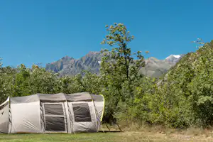 Zelt auf einem Campingplatz mit Bäumen und Bergen im Hintergrund, unter klarem blauen Himmel.