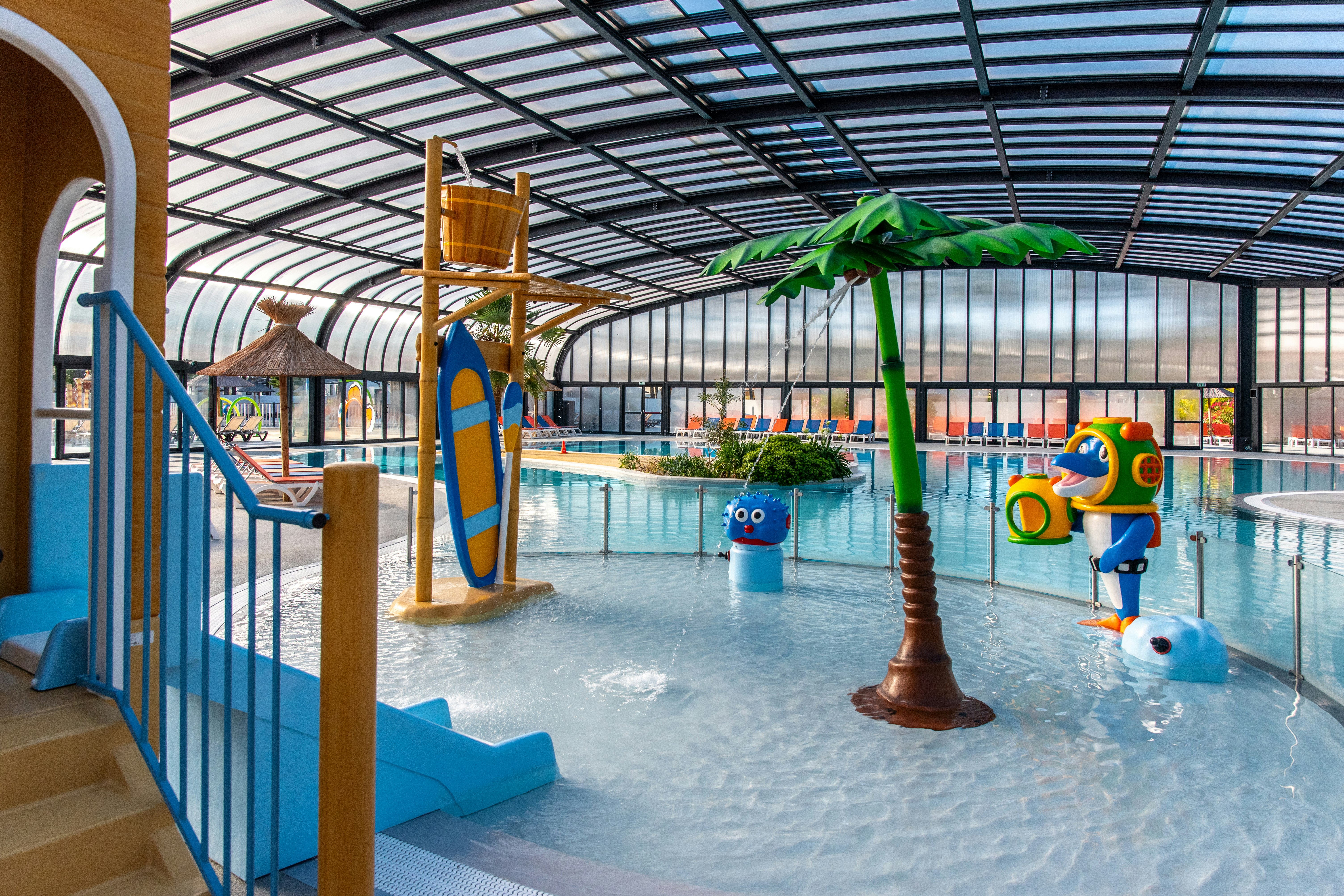 Innenbereich eines Schwimmbads mit überdachtem Becken, Palmen-Deko und bunten Wasserspielzeugen, Liegen und Geländer am Eingang.