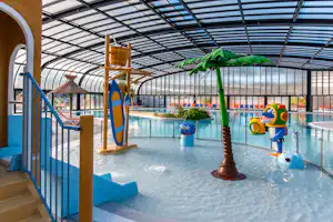 Innenbereich eines Schwimmbads mit überdachtem Becken, Palmen-Deko und bunten Wasserspielzeugen, Liegen und Geländer am Eingang.