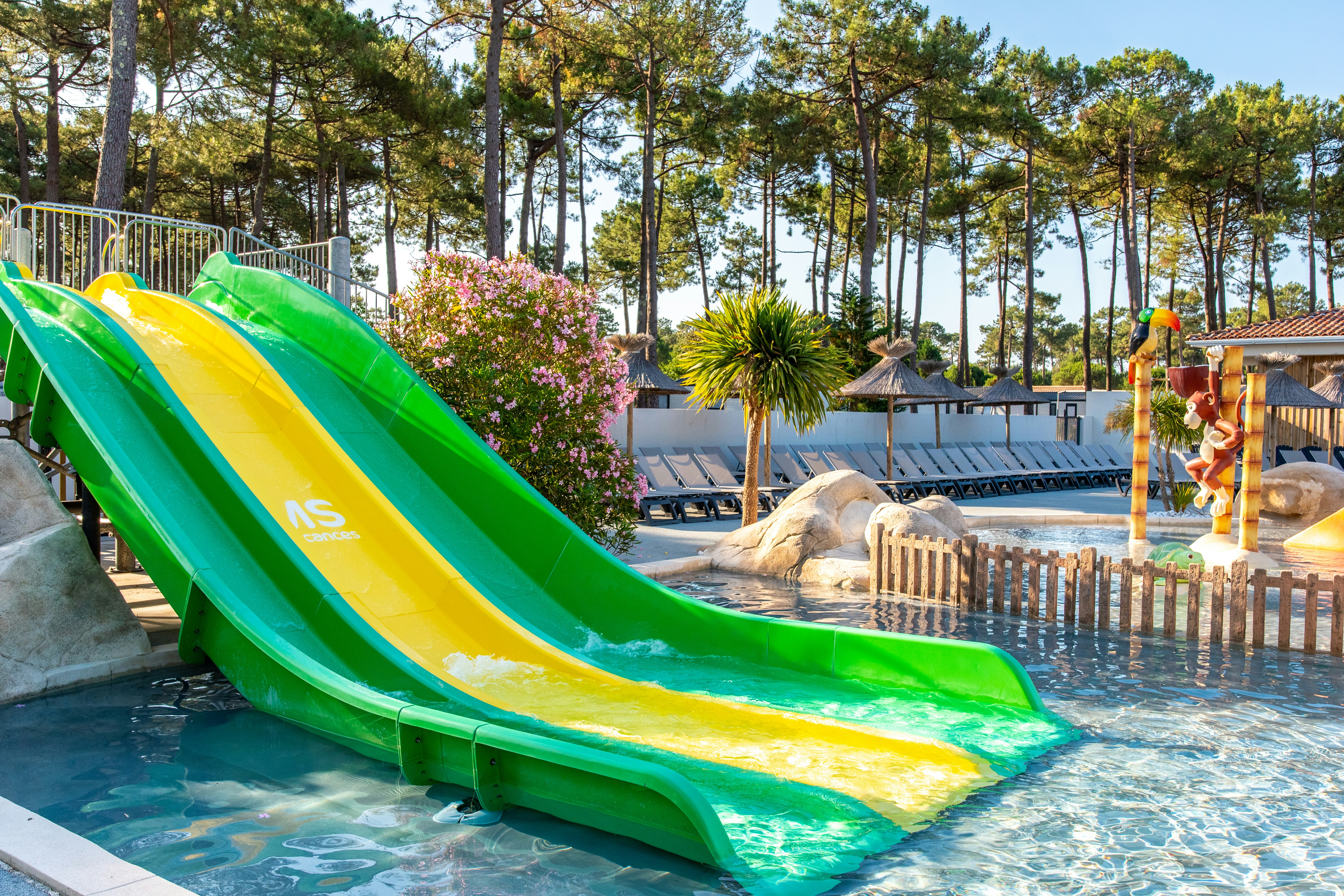 Zwei bunte Wasserrutschen am Pool, daneben ein Liegebereich mit Liegen und Zaun, im Hintergrund Bäume und Hecken.