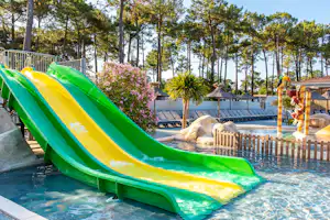 Zwei bunte Wasserrutschen am Pool, daneben ein Liegebereich mit Liegen und Zaun, im Hintergrund Bäume und Hecken.
