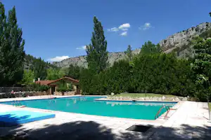 Schwimmbecken im Campingplatz mit Liegen, Sonnenschirmen und Blick auf Bäume und Felsen im Hintergrund.