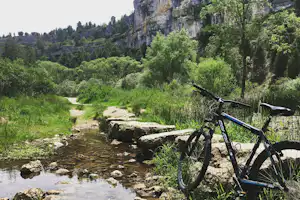 Fahrrad am Bachufer mit Steinen und schmalem Pfad im Grünen, im Hintergrund Felswand und Bäume.