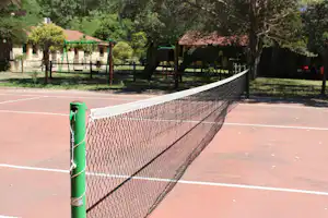 Tennisplatz mit Netz und Pfosten auf dem Campingplatz, im Hintergrund Bäume, Sitzgelegenheiten und ein Spielbereich.