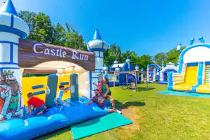 Große blaue Hüpfburg „Castle Run“ mit Rutsche, daneben weitere aufblasbare Spielbereiche auf grünem Rasen.