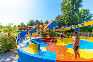Kinder am Camping-Pool mit bunten Wasserrutschen, Spritzwasser und Liegeflächen, im Hintergrund Bäume und Zaun.