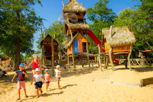 Spielplatz mit Strohhütten, Seilnetz und roter Rutsche auf Sand, umgeben von Bäumen und Holzgeländer.