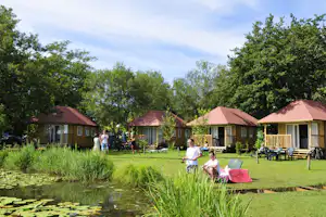 Reihe roter Bungalows auf einem Campingplatz mit Rasen, Wegen und Sitzgelegenheiten, im Vordergrund Kinder im Garten.