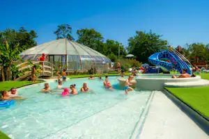 Familienpool mit Wasserrutsche, Kinder planschen im Becken, im Hintergrund ein transparenter Kuppelbau und Bäume.