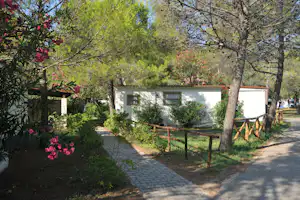 Bungalow im Campinggarten mit gepflastertem Weg, Blumenbeeten, Bäumen und weißen Wänden mit Fenstern.